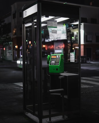 A glass phone booth located on a street corner at night, featuring a bright green payphone. The surrounding environment is dimly lit with a few colored lights from nearby buildings illuminating the scene.