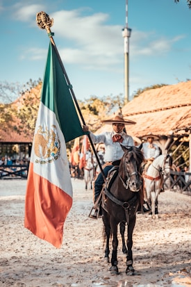 A person wearing traditional attire rides a black horse while holding a large flag, showcasing vibrant colors. Other riders and horses are visible in the background, set against a backdrop of rustic wooden buildings and a clear sky.