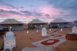 A desert campsite scene featuring several white tents under a scenic dusk sky with vibrant purple and pink hues. The ground is adorned with decorative carpets, and multiple ornate lanterns are placed along the pathways, casting a warm, inviting glow. Comfortable seating with cushions is visible, creating a cozy and exotic atmosphere.