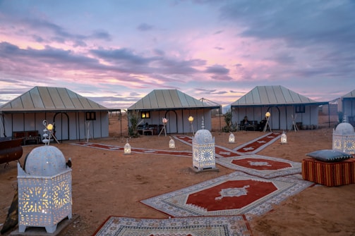 A desert campsite scene featuring several white tents under a scenic dusk sky with vibrant purple and pink hues. The ground is adorned with decorative carpets, and multiple ornate lanterns are placed along the pathways, casting a warm, inviting glow. Comfortable seating with cushions is visible, creating a cozy and exotic atmosphere.