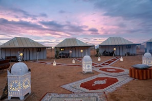 A desert campsite scene featuring several white tents under a scenic dusk sky with vibrant purple and pink hues. The ground is adorned with decorative carpets, and multiple ornate lanterns are placed along the pathways, casting a warm, inviting glow. Comfortable seating with cushions is visible, creating a cozy and exotic atmosphere.