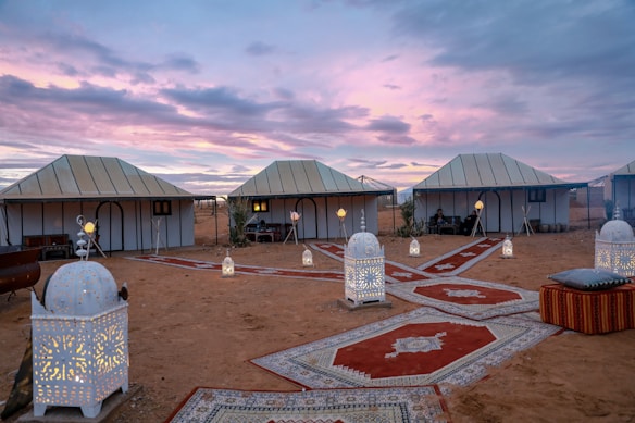 A desert campsite scene featuring several white tents under a scenic dusk sky with vibrant purple and pink hues. The ground is adorned with decorative carpets, and multiple ornate lanterns are placed along the pathways, casting a warm, inviting glow. Comfortable seating with cushions is visible, creating a cozy and exotic atmosphere.
