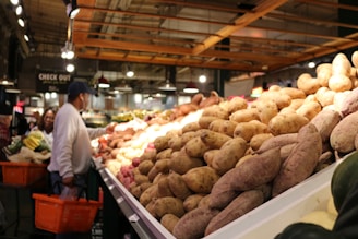 A bustling indoor market featuring a large display of sweet potatoes piled high on a counter. To the left, shoppers with shopping baskets browse the produce section. The setting is well-lit with overhead lights and an industrial-style ceiling.