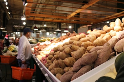 A bustling indoor market featuring a large display of sweet potatoes piled high on a counter. To the left, shoppers with shopping baskets browse the produce section. The setting is well-lit with overhead lights and an industrial-style ceiling.