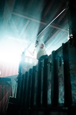 A person is standing on a metal platform, partially obscured by bright light streaming in from the left. The individual is holding onto a railing, and the environment appears industrial, with corrugated metal and beams overhead.