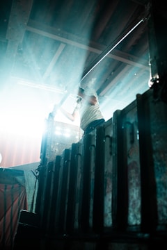 A person is standing on a metal platform, partially obscured by bright light streaming in from the left. The individual is holding onto a railing, and the environment appears industrial, with corrugated metal and beams overhead.