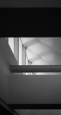 Geometric architectural design featuring intersecting planes and lines. The image is in black and white, emphasizing sharp angles and the play of light and shadow across the surfaces. The structure includes large windows that allow light to cast patterned shadows.