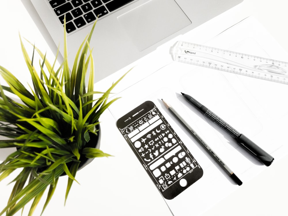 black pen and pencil beside smartphone screen protector beside green plant and laptop computer