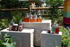 Various jars and bottles of honey placed on white wooden crates outdoors, surrounded by lush greenery and colorful flowers. The containers vary in size and are filled with honey in different shades, from light golden to deep amber. A fence and more vegetation can be seen in the background, suggesting a garden or natural setting.
