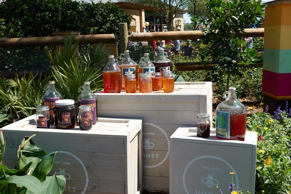 Various jars and bottles of honey placed on white wooden crates outdoors, surrounded by lush greenery and colorful flowers. The containers vary in size and are filled with honey in different shades, from light golden to deep amber. A fence and more vegetation can be seen in the background, suggesting a garden or natural setting.