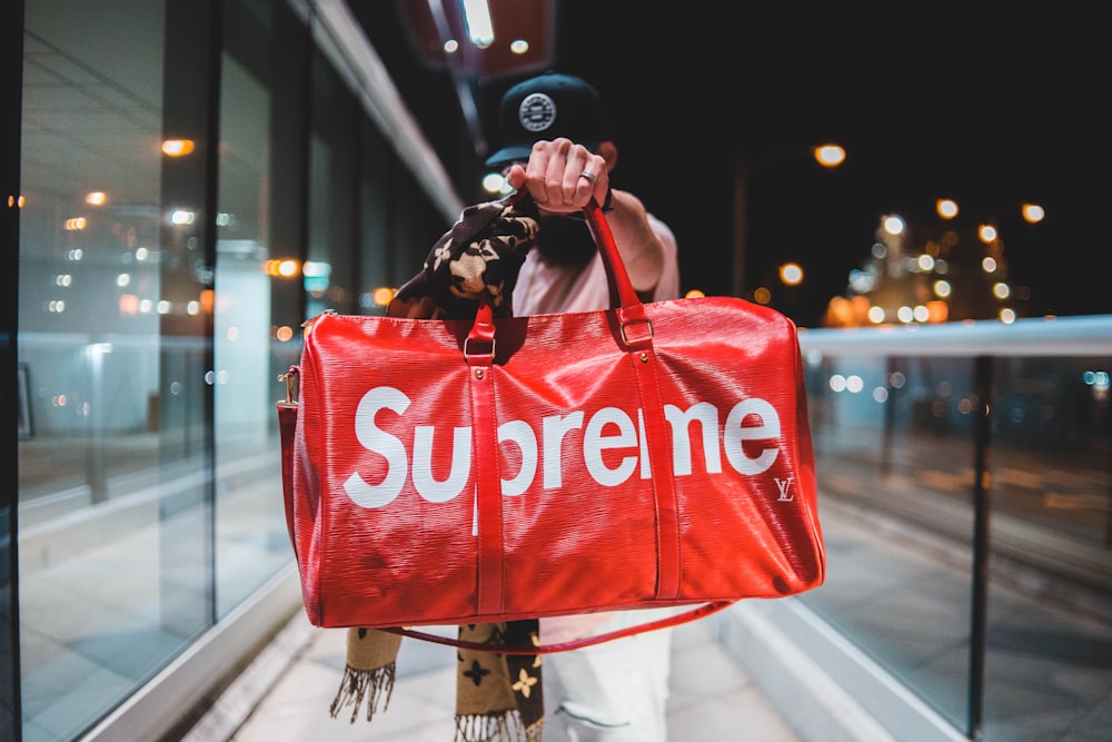 person holding red and white Supreme leather duffel bag outdoor