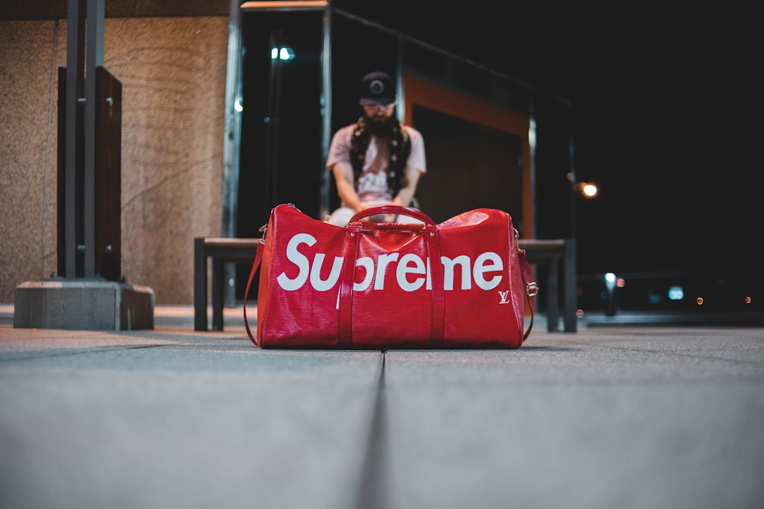 man sitting on brown bench in front of red and white leather Supreme duffel bag