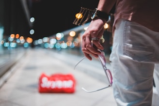 A hand is holding sunglasses and wearing a watch and rings, with a red bag in the distance on a walkway lined by glass railings and blurred lights in the background.