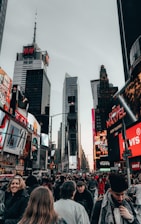 A bustling cityscape filled with a large crowd of people, surrounded by towering buildings featuring bright electronic billboards and advertisements. The scene captures the busy and vibrant essence of an urban environment, likely a famous city plaza.