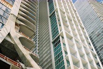 A perspective view of modern high-rise buildings with detailed architectural design. The structures feature a combination of concrete and glass elements, with numerous balconies and a grid-like pattern on the surfaces.