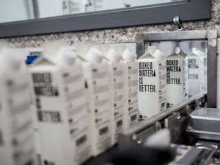 A row of boxed water cartons with the text 'BOXED WATER IS BETTER' on them, positioned on a conveyor belt in what appears to be a manufacturing or packaging facility. The background includes a mirror and a wall with a speckled texture.