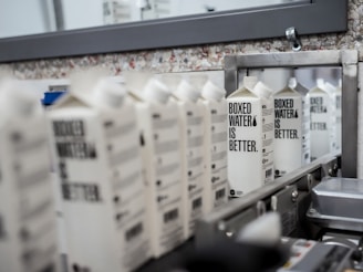 A row of boxed water cartons with the text 'BOXED WATER IS BETTER' on them, positioned on a conveyor belt in what appears to be a manufacturing or packaging facility. The background includes a mirror and a wall with a speckled texture.