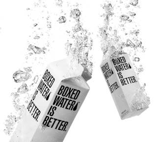 Two cartons of Boxed Water are surrounded by splashing water droplets, creating a dynamic and energetic scene. The cartons have bold black text on a white background.