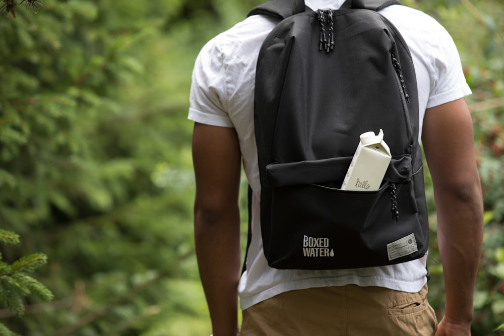 A man standing in the outdoors with a black backpack and a white Boxed Water box sticking out