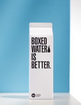 A white carton prominently features the text 'BOXED WATER IS BETTER.' against a simple blue background. The carton has a clean, minimalistic design with bold black lettering.