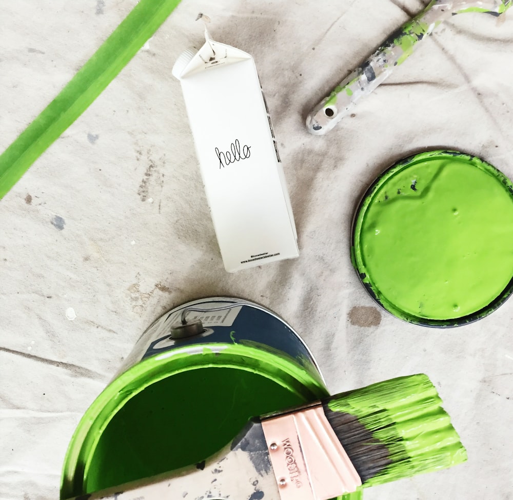 Una lata de pintura verde y un pincel junto a un cartón de agua en caja