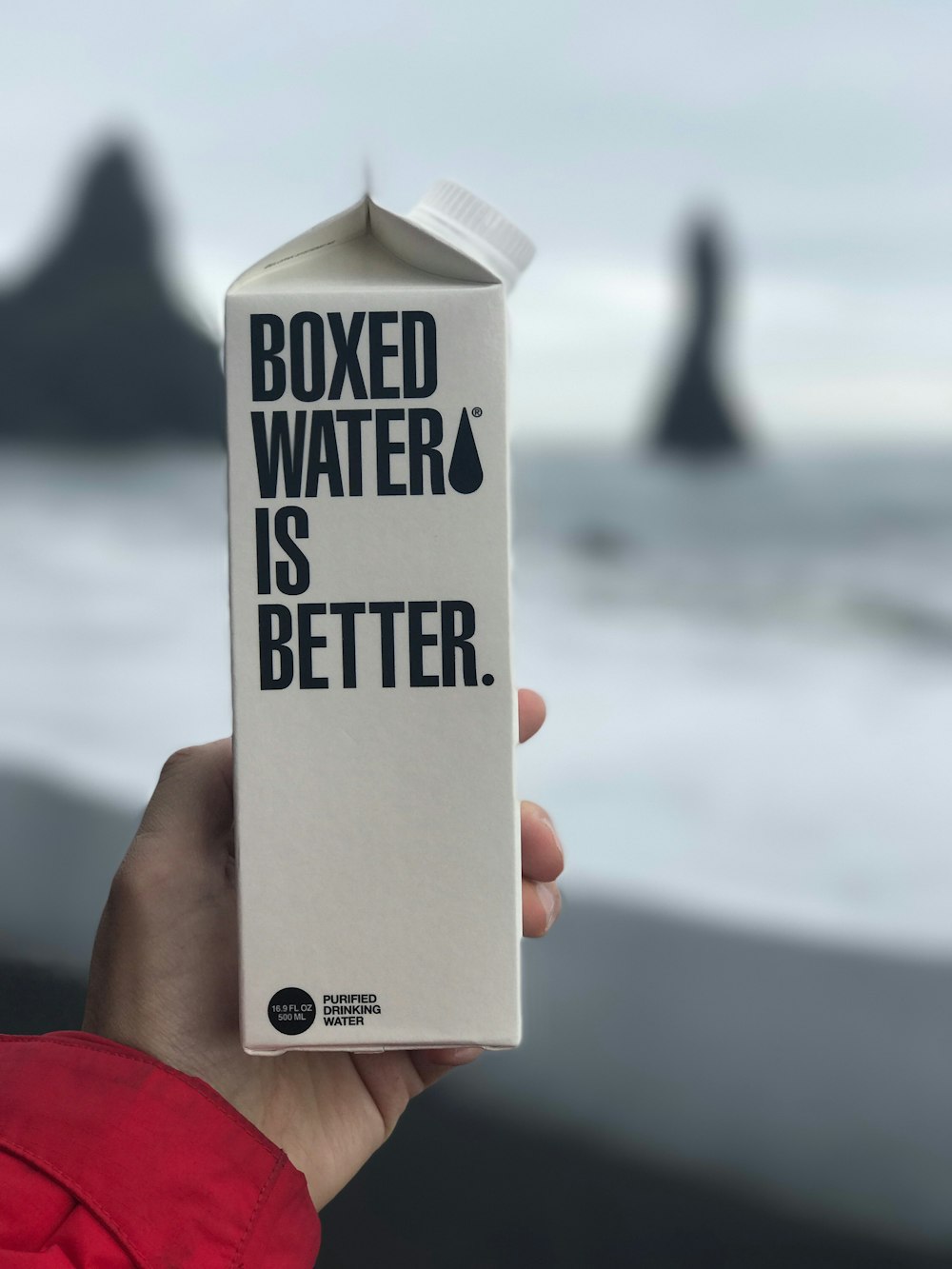 A Boxed Water carton is held in front of an Icelandic beach