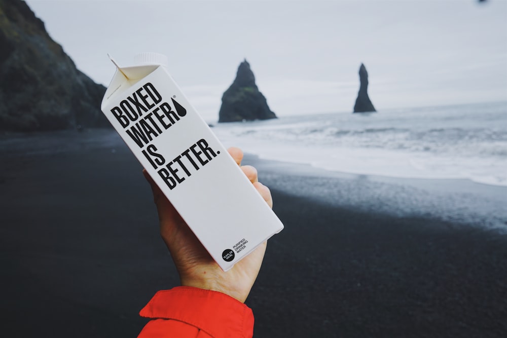 A Boxed Water carton is held by a person in front of an Icelandic beach