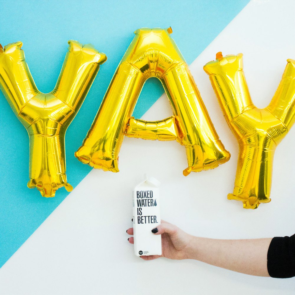 A person holding a Boxed Water carton in front of balloons spelling YAY