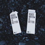 Two cartons labeled 'Boxed Water Is Better' rest on a bed of smooth, dark stones. The cartons are white and feature bold black text, emphasizing an eco-friendly message.
