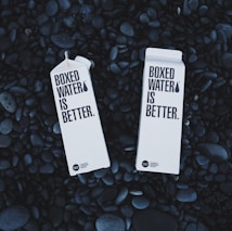 Two cartons labeled 'Boxed Water Is Better' rest on a bed of smooth, dark stones. The cartons are white and feature bold black text, emphasizing an eco-friendly message.