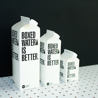 A set of white cartons with black text are arranged in a row on a white polka-dotted surface against a black background. Each carton is labeled 'Boxed Water is Better,' and they vary in size from large to small, creating a sense of depth and scale.
