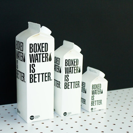 A set of white cartons with black text are arranged in a row on a white polka-dotted surface against a black background. Each carton is labeled 'Boxed Water is Better,' and they vary in size from large to small, creating a sense of depth and scale.