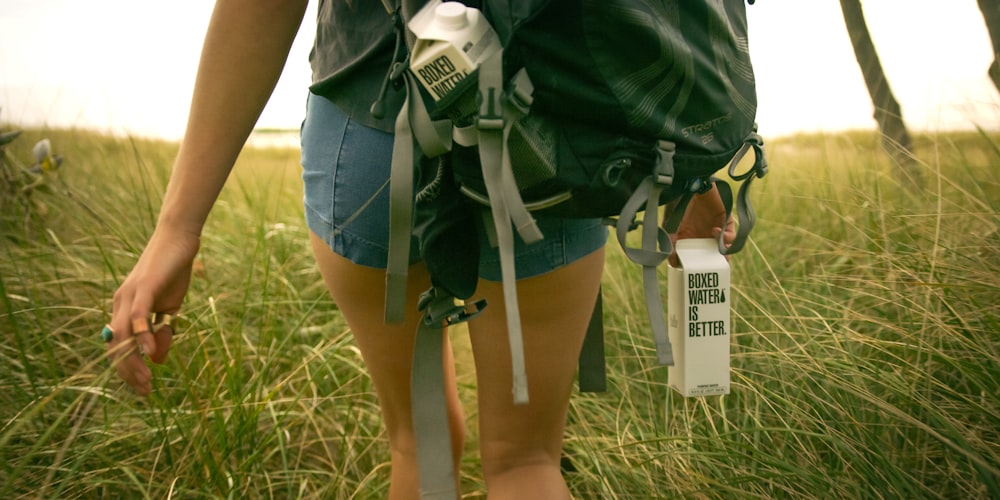 A woman walking in a field holding a Boxed Water carton
