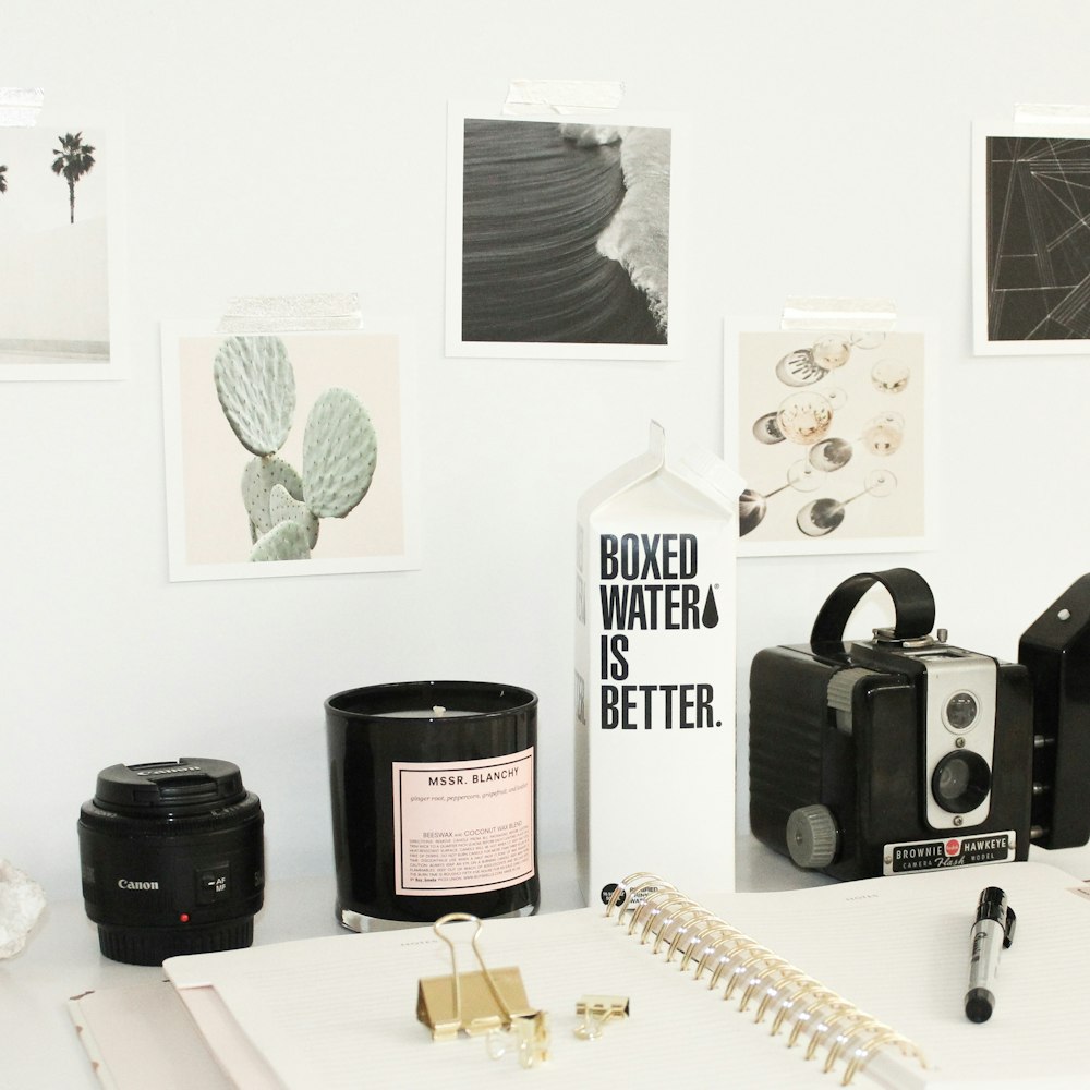 A black camera lens beside a white spring notebook and Boxed Water carton