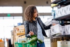 A person with curly hair wearing a leather jacket and jeans shops in a store aisle. They are holding a box labeled 'Sustainability Matters' and have a green shopping basket with fresh produce, including leafy greens.