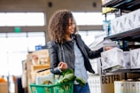 A person with curly hair wearing a leather jacket and jeans shops in a store aisle. They are holding a box labeled 'Sustainability Matters' and have a green shopping basket with fresh produce, including leafy greens.