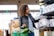 A person with curly hair wearing a leather jacket and jeans shops in a store aisle. They are holding a box labeled 'Sustainability Matters' and have a green shopping basket with fresh produce, including leafy greens.