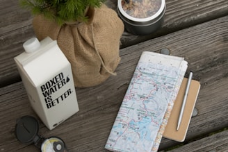 A wooden table surface holds a carton of boxed water with the text 'BOXED WATER IS BETTER' printed on it. Beside it, a wrapped plant in burlap and a tin of mixed nuts or trail mix are arranged. A folded map, partially showing an area called Yellowstone National Park, lies next to a pen and a small notebook. A compass is also present, indicating a theme of outdoor adventure or travel preparation.