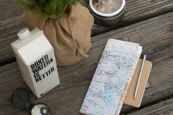 A wooden table surface holds a carton of boxed water with the text 'BOXED WATER IS BETTER' printed on it. Beside it, a wrapped plant in burlap and a tin of mixed nuts or trail mix are arranged. A folded map, partially showing an area called Yellowstone National Park, lies next to a pen and a small notebook. A compass is also present, indicating a theme of outdoor adventure or travel preparation.