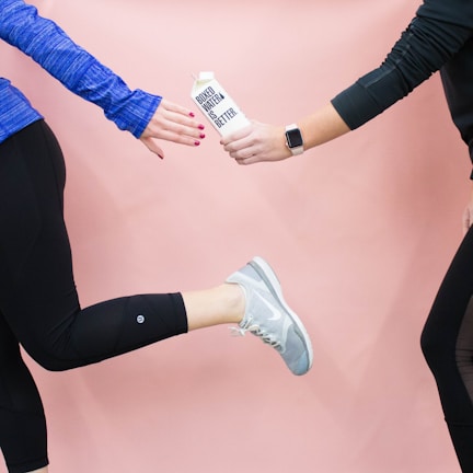 Two individuals are engaged in a handoff of a carton labeled 'Boxed Water is Better.' One person is dressed in a blue long-sleeved top and black leggings, holding the carton, while the other, visible only in part, is wearing a black top and has a smartwatch on their wrist. The background is a soft pink color.