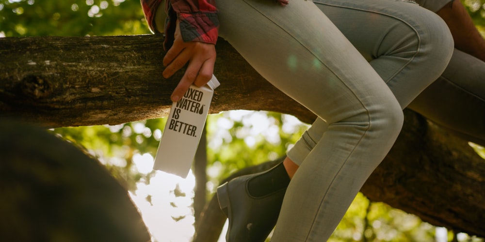 A person sits on a tree branch holding a Boxed Water carton