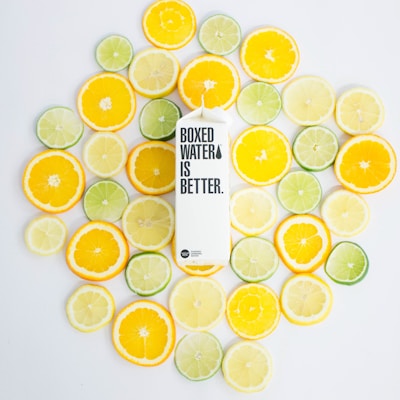 A white carton labeled 'Boxed Water is Better' is surrounded by neatly arranged slices of citrus fruits, including oranges, lemons, and limes, on a light background.