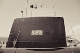 A large submarine with the number S22 painted on its side is docked at a port. Multiple antennas and periscopes protrude from the top. A person stands near the base of the submarine, looking upward. The scene appears to be in a maritime environment with additional ships visible in the background.