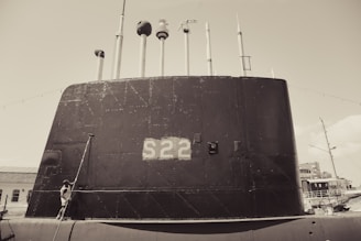 A large submarine with the number S22 painted on its side is docked at a port. Multiple antennas and periscopes protrude from the top. A person stands near the base of the submarine, looking upward. The scene appears to be in a maritime environment with additional ships visible in the background.