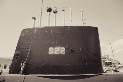 A large submarine with the number S22 painted on its side is docked at a port. Multiple antennas and periscopes protrude from the top. A person stands near the base of the submarine, looking upward. The scene appears to be in a maritime environment with additional ships visible in the background.