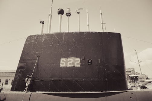 A large submarine with the number S22 painted on its side is docked at a port. Multiple antennas and periscopes protrude from the top. A person stands near the base of the submarine, looking upward. The scene appears to be in a maritime environment with additional ships visible in the background.