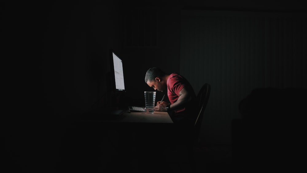 赤いシャツを着た男が、パソコンのモニターの前で机にもたれかかりながら書き物をしている