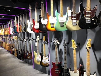 A variety of electric and acoustic guitars are displayed along a wall in a well-lit showroom. The guitars are positioned on two rows of sturdy mounts, showcasing a wide range of colors and finishes. The lighting accentuates the vibrant hues and wooden textures, with some guitars featuring metallic or glossy finishes.