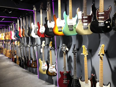 A variety of electric and acoustic guitars are displayed along a wall in a well-lit showroom. The guitars are positioned on two rows of sturdy mounts, showcasing a wide range of colors and finishes. The lighting accentuates the vibrant hues and wooden textures, with some guitars featuring metallic or glossy finishes.