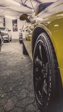 A close-up of a car tire and wheel, with a bold yellow car body reflecting ambient light. The image is taken in a garage with several cars parked in the background. The ceiling has an industrial look with metal beams and white fluorescent lights.